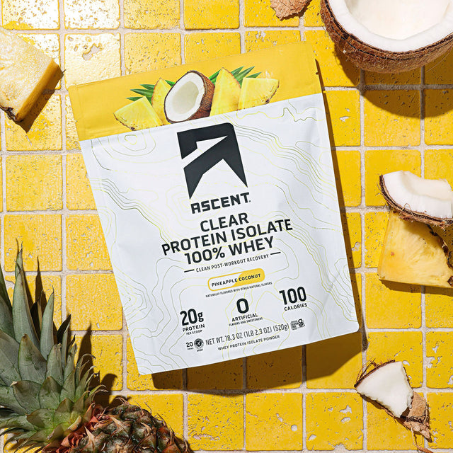 A bag of Pineapple Coconut Clear Whey laying on a yellow tile surface surrounded by cracked coconuts and sliced pineapple.