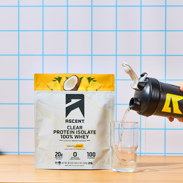 A bag of Pineapple Coconut Clear Whey Protein next to a glass being filled by a shaker bottle with Pineapple Coconut Clear Whey mixed with water.