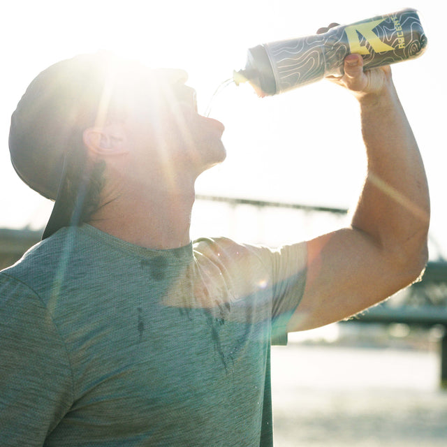 A man in a gray t-shirt drinking from a black and gold patterned water bottle, captured in bright backlit sun with lens flare. split_row_banner_2