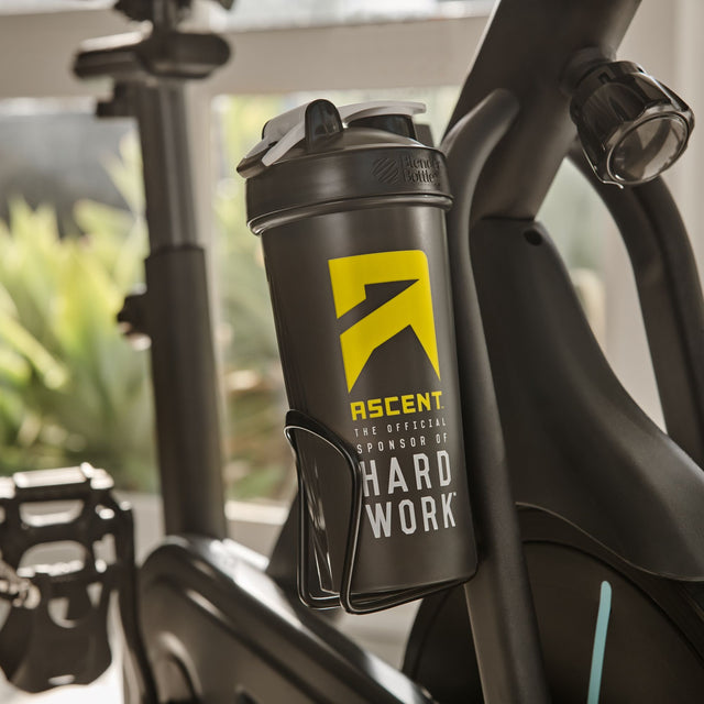 Black shaker bottle with yellow and white text attached to a bicycle frame, featuring the 'Ascent' brand saying Ascent: the official sponsor of hard work split_row_banner_2