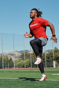 Person in red 'Ascent' shirt running on a sports field with mountains in the background