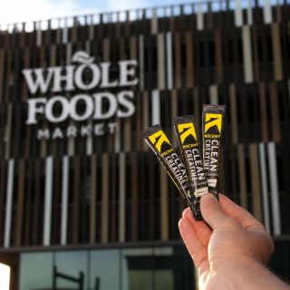 A hand holds three slim black and yellow Ascent Clean Creatine sticks in front of a blurred Whole Foods Market storefront.