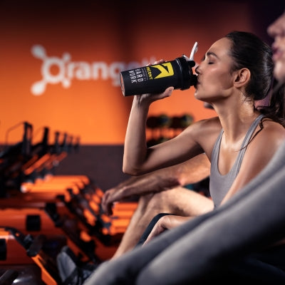 ascent protein athlete drinking from a protein powder blender bottle with the orange theory logo in the background