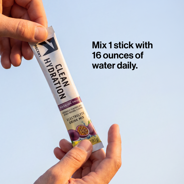 A person holds a single stick of Ascent Clean Hydration Passionfruit electrolyte drink mix against a light blue sky background, with text on the right that reads, 'Mix 1 stick with 16 ounces of water daily.'.