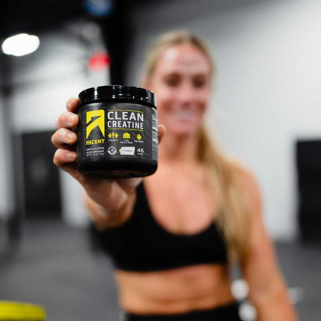 Woman holding a container of 'Ascent Clean Creatine' powder in a gym setting.