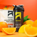 A bag of Orange Mango Clear Whey Protein sitting next to a shaker bottle and sliced mangos and oranges.