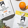 A bag of Orange Mango Clear Whey protein powder laying next to a bag of Pineapple Coconut Clear Whey protein powder as well as a pineapple, orange, and coconut.
