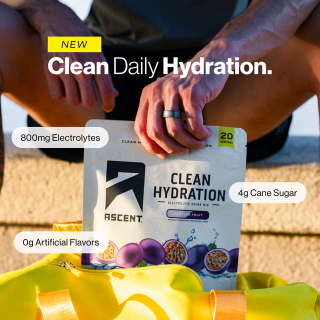 A person outdoors holds a packet of ASCENT Clean Hydration electrolyte drink mix. The package is white with black text and fruit graphics, with callouts reading 800mg Electrolytes, 4g Cane Sugar, and 0g Artificial Flavors.