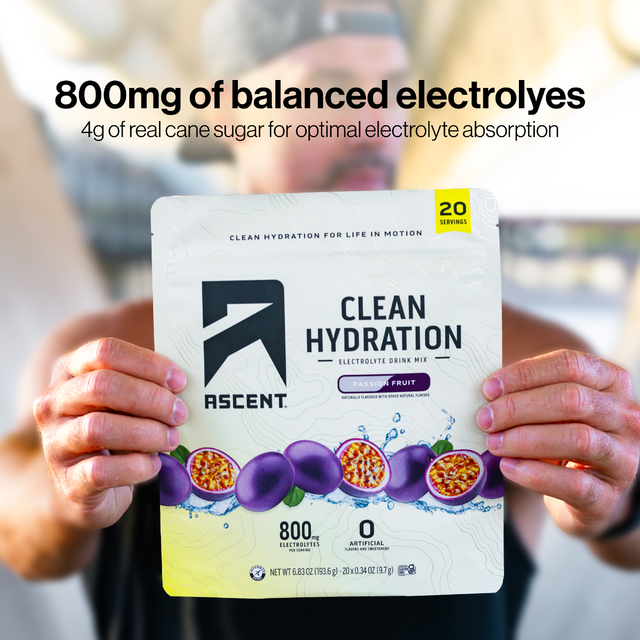A person holds up a packet of Ascent Clean Hydration Electrolyte Drink Mix in Passion Fruit flavor. The packet is white with purple passion fruit graphics and black text.