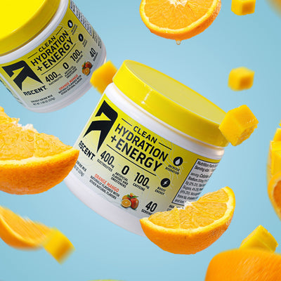 Two containers of 'Clean Hydration + Energy' supplement surrounded by flying oranges and mangoes on a blue background.