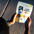 A person holds an Ascent Clean Hydration electrolyte drink pouch outdoors on a sunny day. The white pouch features black branding and illustrations of strawberries and citrus slices, with the person's hands and head visible in the foreground. split_row_banner_1