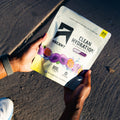 A person holds an Ascent Clean Hydration electrolyte powder pouch in their hands, photographed from above on an asphalt surface with strong sunlight and shadows. split_row_banner_1
