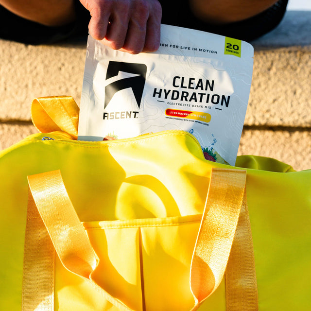 A person's hand holding an ASSENT Clean Hydration electrolyte powder bag above a bright yellow tote bag outdoors on a sunny day. split_row_banner_2