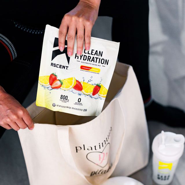 A person holds a bag of ASCENT Clean Hydration powder and pours it into a large cream tote bag printed with 'platinum pilates' and a heart logo. A white shaker bottle with a yellow logo sits nearby on a gray surface. split_row_banner_2