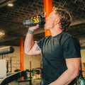 A man in a gym drinking from a black and yellow shaker bottle. 
split_row_banner_2