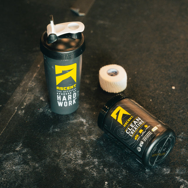 Ascent Clean Creating 100 serving container laying on the ground next to an Ascent shaker bottle