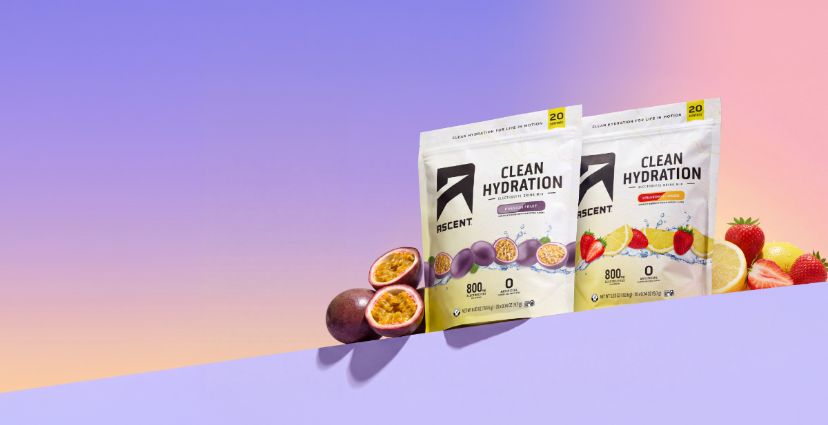 A bag of passion fruit flavored Ascent Clean Hydration and a bag of Strawberry Lemonade Ascent Clean Hydration sitting on a platform next to passion fruits, strawberries, and lemons in front of a purple and pink gradient background.