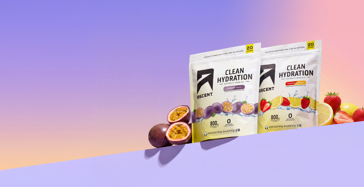 Two stand-up pouches of ASCENT Clean Hydration drink mix are displayed on a stylized surface against a pastel gradient background. The pouches are shown with fruit slices and whole fruits like passion fruit, strawberries, and lemons arranged around them.
