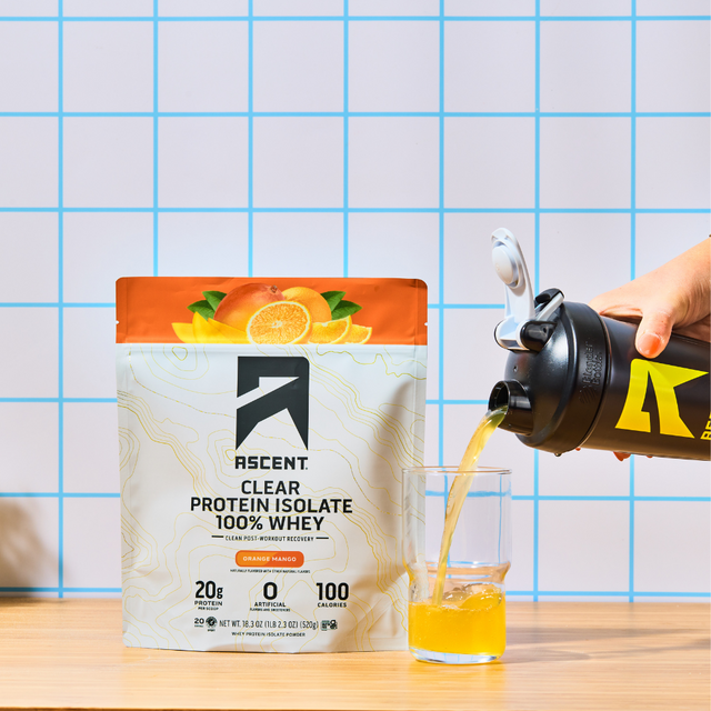 A bag of Orange Mango Clear Whey protein powder sitting next to a glass being filled with the orange mango clear whey mixed with water from a shaker bottle.
