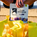 A person holds an ASCENT Clean Hydration electrolyte drink mix pouch above a bright yellow duffel bag outdoors on stone steps. split_row_banner_2