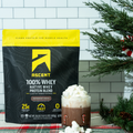 Ascent protein blend bag in a holiday scene with peppermint mocha drink in a glass with candy canes on it and pine tree next to it