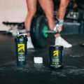 Pre-workout supplement containers on a gym floor with a person lifting weights.