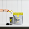 A hand holding a plastic scoop and pouring Micellar Casein powder into a shaker bottle next to a bag of Vanilla Bean Micellar Casein.