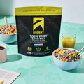 Ascent 100% Fruity Cereal Whey Protein Blend package on a table with bowls of cereal and drinks.