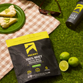 Ascent 100% Key Lime Pie Whey Protein Powder package on grass with lime, pie, and water bottle.