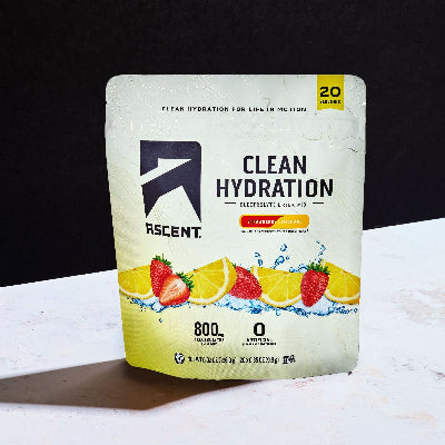 A stand-up pouch of ASCENT Clean Hydration Electrolyte Drink Mix, strawberry lemonade flavor, standing on a white marble-like surface against a black background.