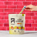A person's hand inserts a single Ascent Hydration stick pack into a large stand-up pouch of Ascent Clean Hydration electrolyte drink mix. The pouch is cream and yellow with illustrations of strawberries and lemons, set against a bright pink tiled wall and a white marble surface.