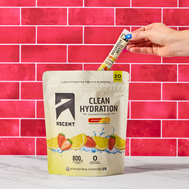A person's hand inserts a single Ascent Hydration stick pack into a large stand-up pouch of Ascent Clean Hydration electrolyte drink mix. The pouch is cream and yellow with illustrations of strawberries and lemons, set against a bright pink tiled wall and a white marble surface.