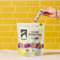 A hand inserts a single-serve stick packet of Ascent Clean Hydration into a large 20-count pouch of the same product, set against a yellow subway tile wall and marble countertop.