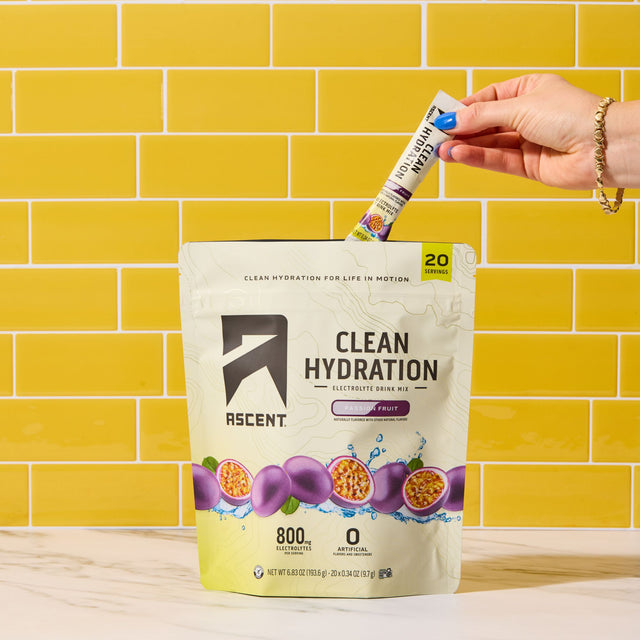 A hand inserts a single-serve stick packet of Ascent Clean Hydration into a large 20-count pouch of the same product, set against a yellow subway tile wall and marble countertop.