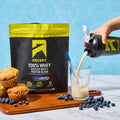 A bag of Ascent 100% Whey Native Whey Protein Blend in Blueberry Muffin flavor stands on a wooden board with blueberries and muffins, while a hand pours a creamy beige shake into a clear glass from a black and yellow shaker bottle. The background is a bright blue geometric wall.