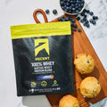 A bag of Ascent 100% Whey Native Whey Protein Blend in Blueberry Muffin flavor, styled on a wooden cutting board with blueberries and muffins on a white marble surface.