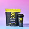 A bag of Ascent 100% Whey Native Whey Protein Blend in Blueberry Muffin flavor next to a matching black shaker bottle with a yellow logo, set against a pink-to-blue gradient studio background.
