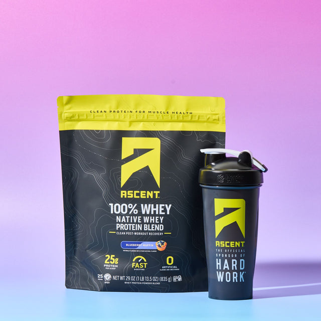A bag of Ascent 100% Whey Native Whey Protein Blend in Blueberry Muffin flavor next to a matching black shaker bottle with a yellow logo, set against a pink-to-blue gradient studio background.