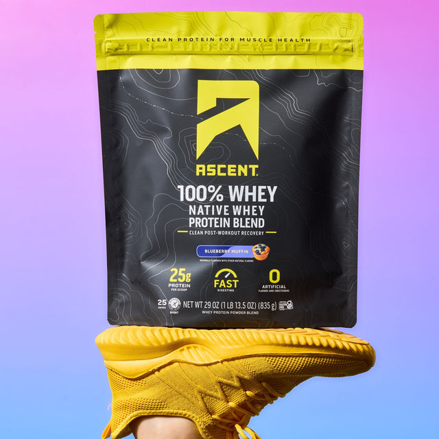 A bag of Ascent 100% Whey Native Whey Protein Blend in Blueberry Muffin flavor, held up by a person's foot wearing a yellow athletic shoe, against a purple and blue gradient background. split_row_banner_1