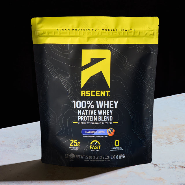 A bag of Ascent 100% Whey Native Whey Protein Blend in Blueberry Muffin flavor, standing on a light marble surface against a black background.