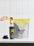 Person pouring protein powder into a shaker cup with a protein package labeled '100% Casein Micellar Isolate' on a white countertop.