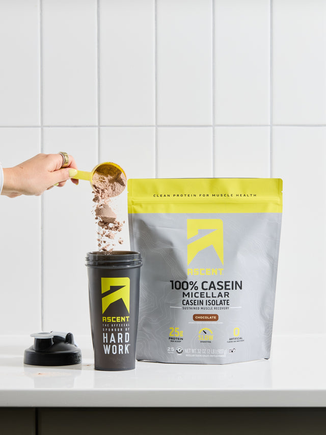 Person pouring protein powder into a shaker cup with a protein package labeled '100% Casein Micellar Isolate' on a white countertop.