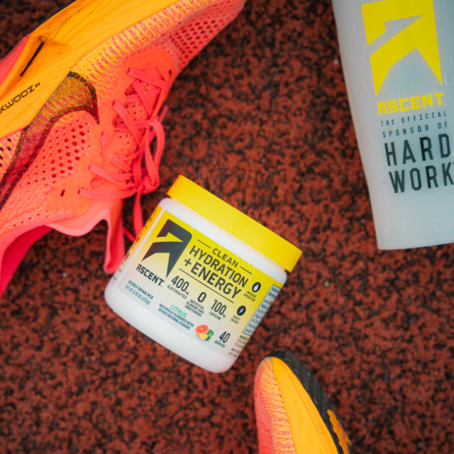 jar of clean hydration + energy powder on a track & field track with shoes and cup next to it
